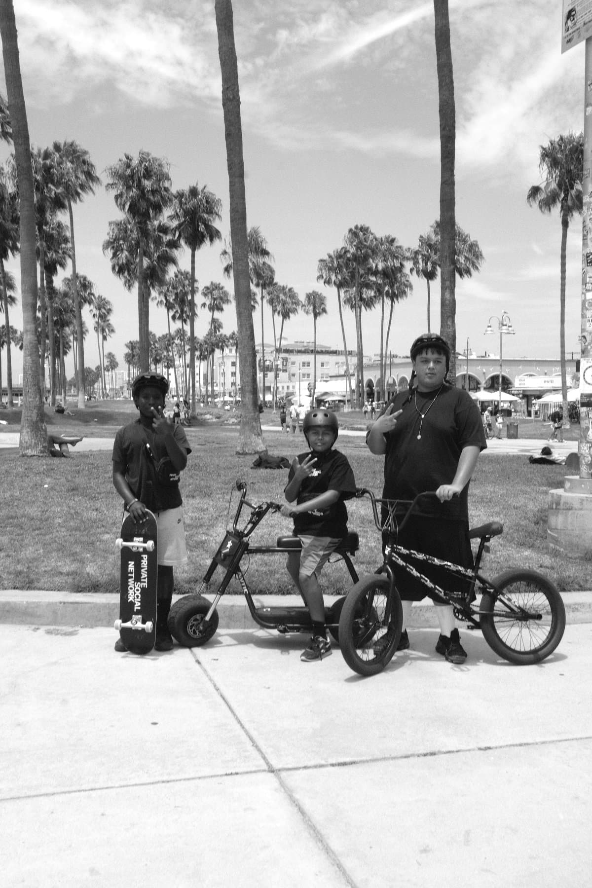 Three young men with a skateboard, electric bike, and BMX at a palm lined park.