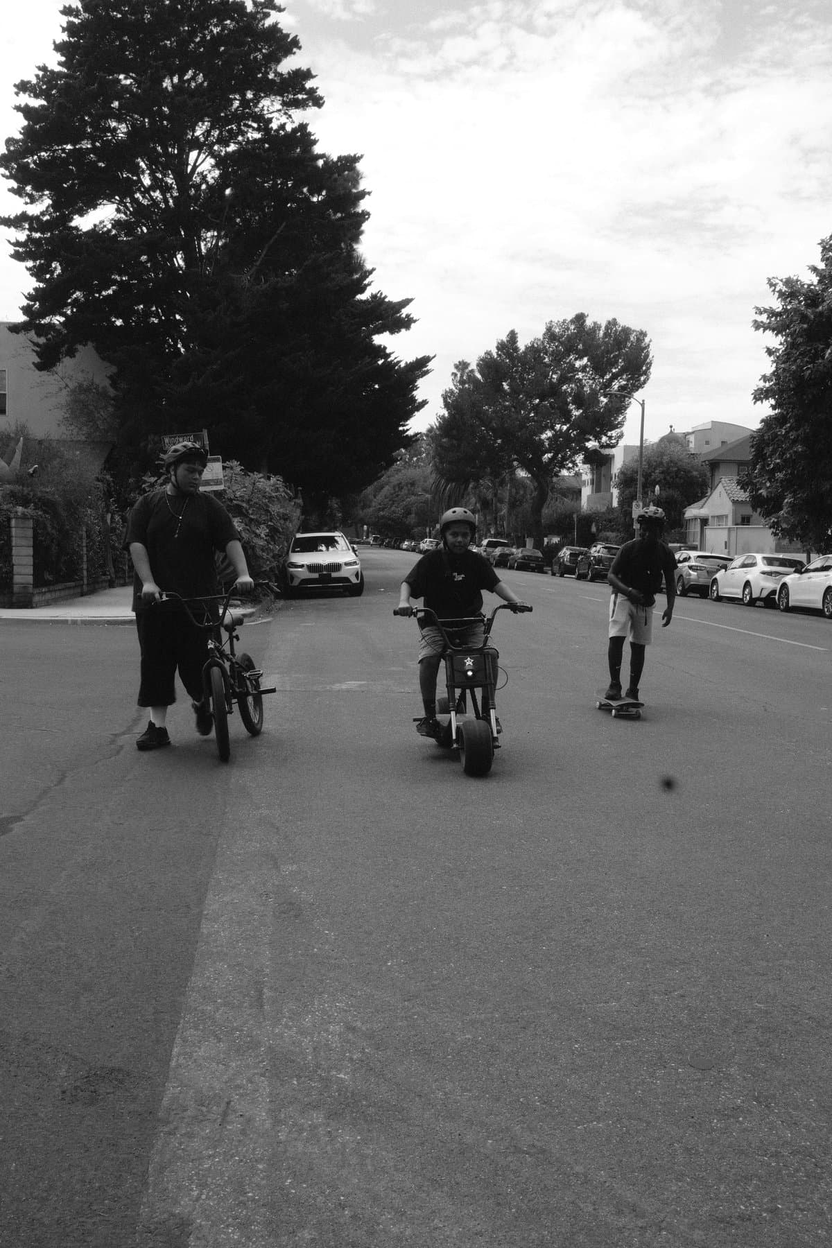 Three young men riding a BMX, an electric bike, and a skateboard down a residential street.